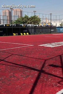 A sports court with a red playing surface and white lines, surrounded by a black mesh fence. In the background, tall buildings are visible, along with some trees and a few yellow chairs positioned near the fence.